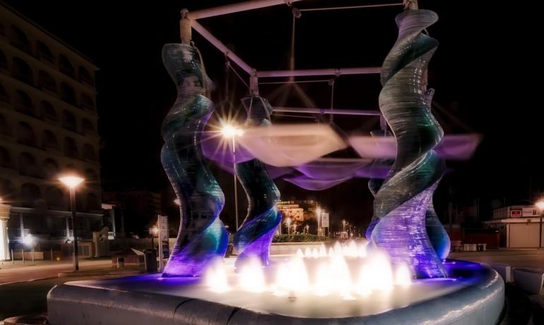 Fontana luminosa con sculture a spirale in una piazza notturna.