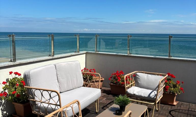 Terrazza soleggiata con vista mare, arredi eleganti e fiori rossi vibranti.